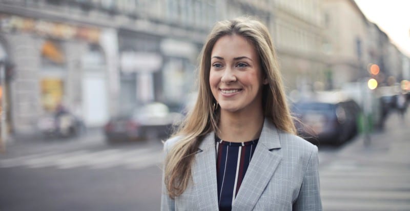 Smiling blonde-haired woman wearing a business blazer and blouse while standing by a pedestrian crosswalk in a city