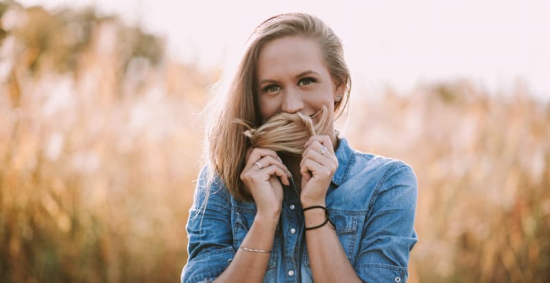 Blonde young woman wears a denim shirt in a golden field while covering her mouth with her hair due to fuzzy teeth