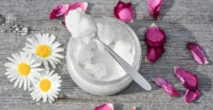 Aerial view of coconut oil in a clear jar with a spoon surrounded by 3 daisies and pink rose petals on a gray counter