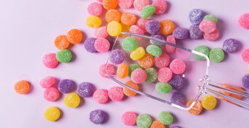 Aerial view of yellow, pink, orange, green, and purple sugary and sticky gumdrops on a lavender counter with a clear scooper
