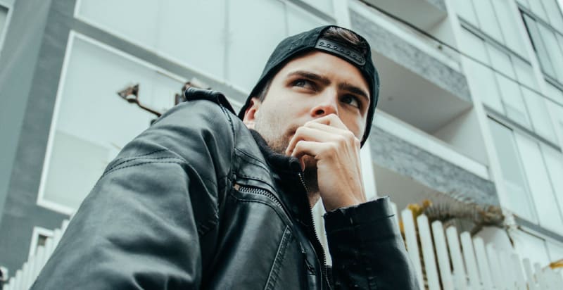 Young man wearing a black leather jacket and backwards baseball cap anxiously covers his mouth due to bad breath from tonsil stones