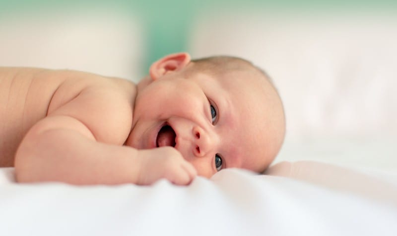 Little baby lies on his belly on white sheets and smiles, showing his gums