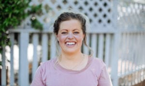 Blonde woman wearing a pink shirt smiles in front of a white picket fence as she thinks about her interesting teeth