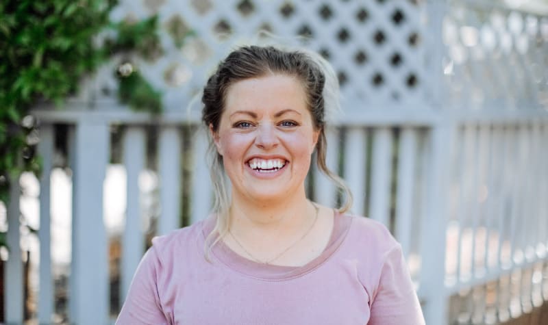 Blonde woman wearing a pink shirt smiles in front of a white picket fence as she thinks about her interesting teeth