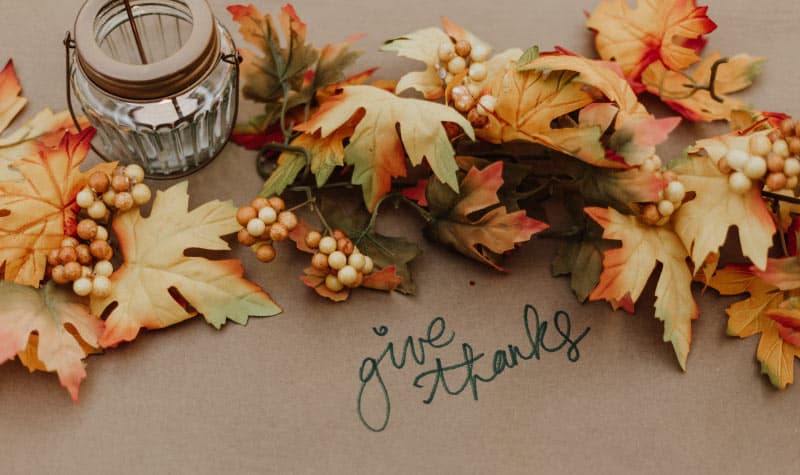 A cluster of fall leaves next to a candle on brown paper with the words "give thanks" to celebrate Thanksgiving